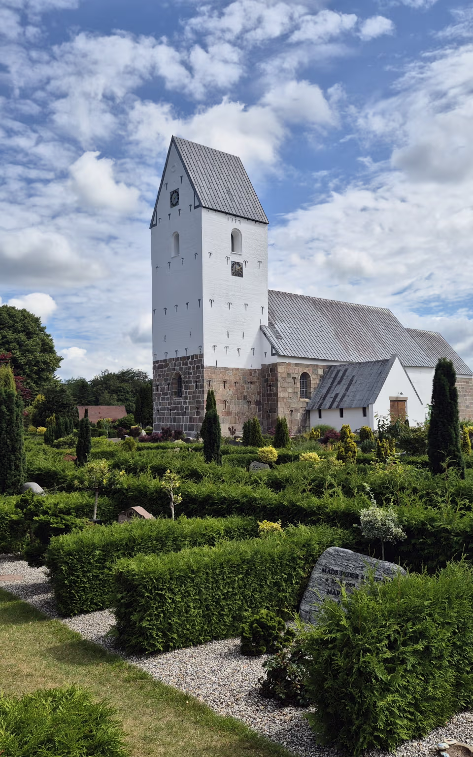 Ølgod Kirke med hvidt tårn bag grønne gravsteder og hække på kirkegården