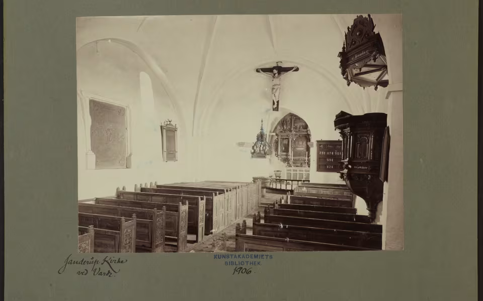 Janderup Kirkes indre i 1906. Hvælvingerne er endnu hvidkalkede, og de middelalderlige kalkmalerier ligger skjult under kalken. Først i 1936 blev de fremdraget. Bemærk også de gamle kirkestole, prædikestolen og den store altertavle fra 1645. Foto: Kunstakademiets Bibliotek, 1906.