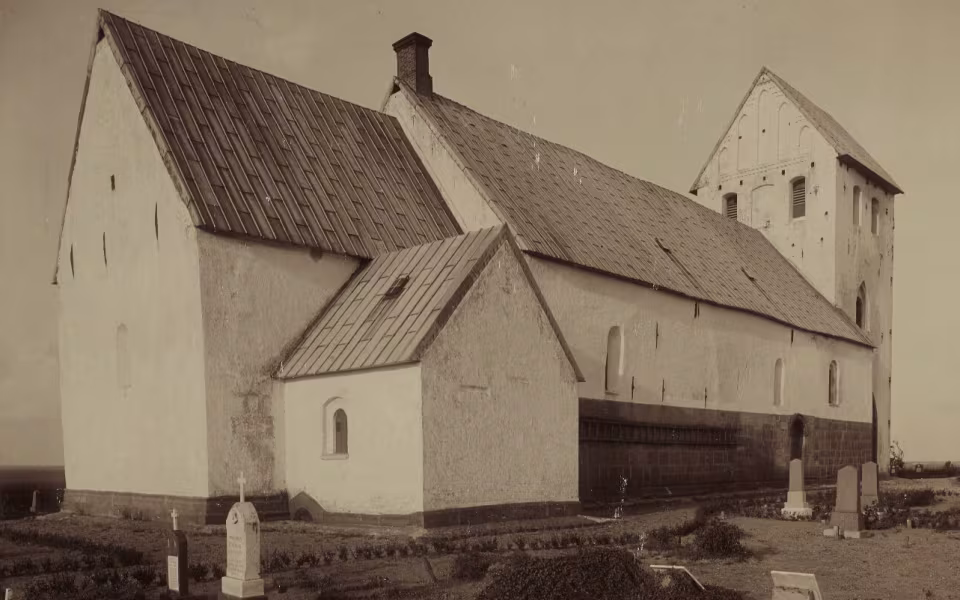 Janderup Kirke og kirkegården i 1906, tre år før den voldsomme stormflod, hvor omkring halvdelen af gravene sank sammen, og gravsten væltede. På kirkens sydside ses det gamle våbenhus, som blev revet ned i 1916. Foto: Kunstakademiets Bibliotek, 1906.