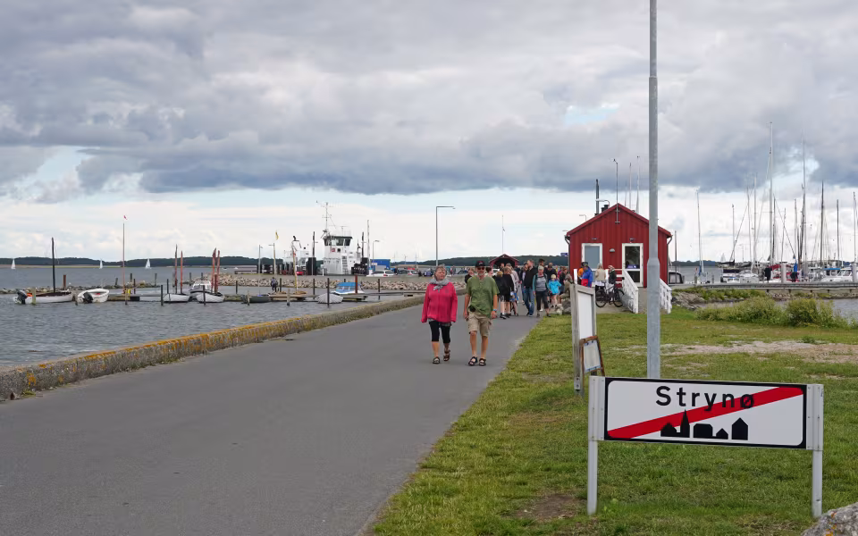 Havn på Strynø med sejlbåde, færgebygninger, gående folk og Strynø-skilt