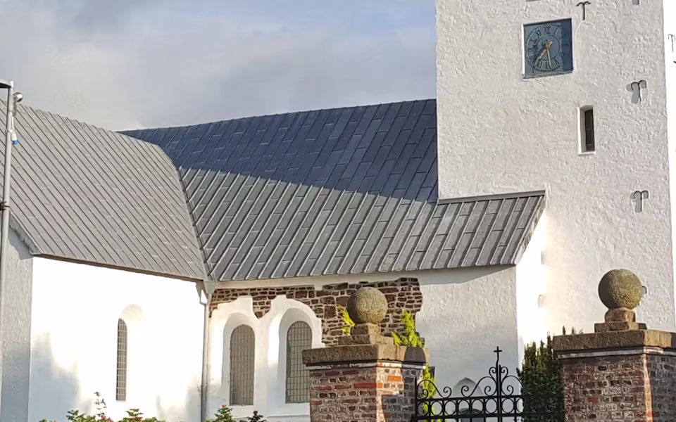 Hvid kirke med tårn, kirkegårdsmur og grøn hæk under klar himmel