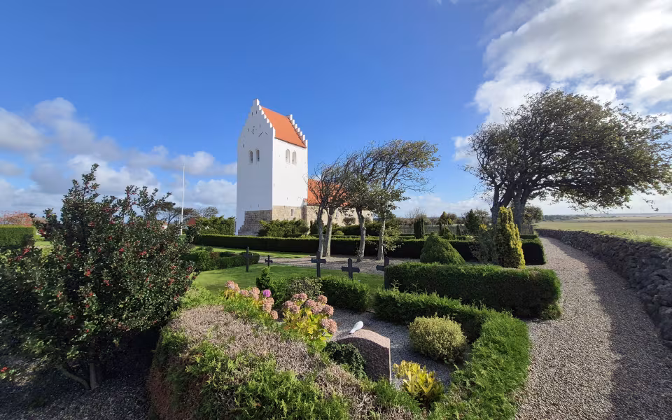 Tømmerby Kirke med hvidt trappetårn og rødt tegltag bag kirkegårdens hække og gravkors