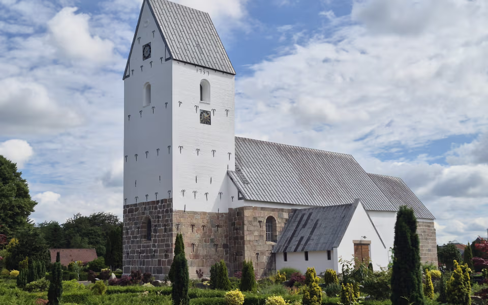Ølgod Kirke med hvidt tårn og grøn kirkegård under skyet himmel