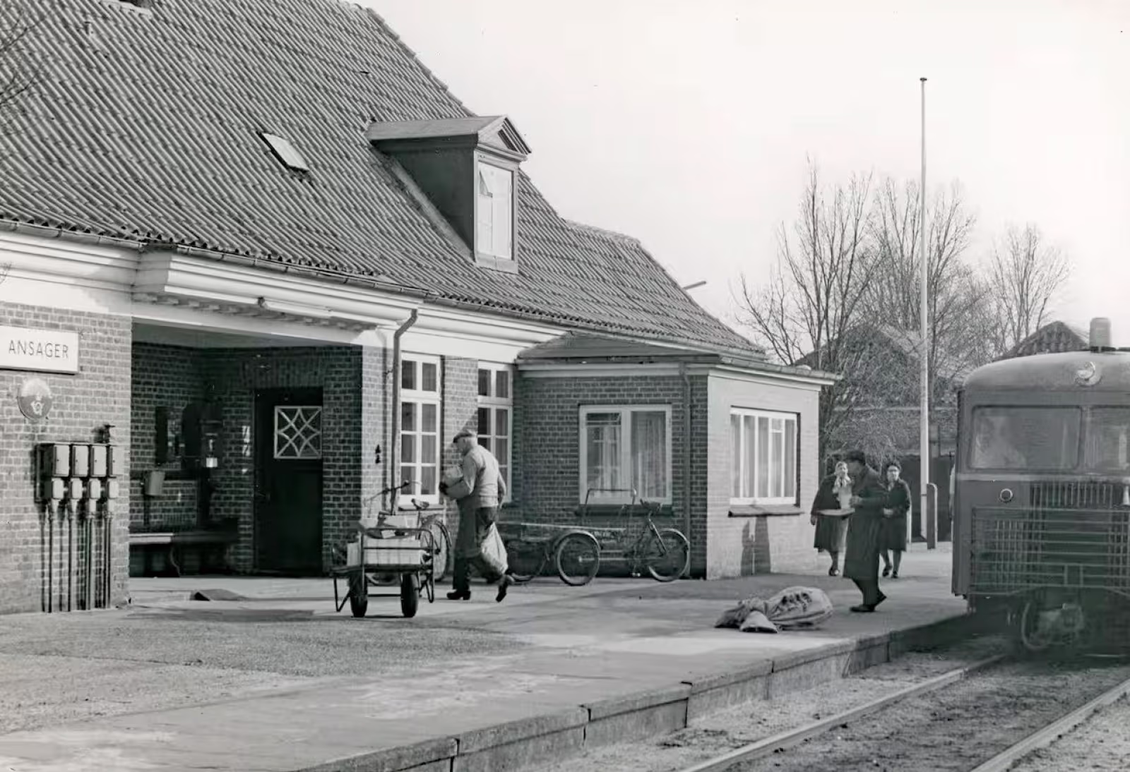 Foto fra togudstillingen: Persontoget fra Grindsted er netop ankommet til Ansager Station, hvor post og pakker bliver fordelt. I forgrunden ses Hans S. Nissen, kaldet “Pakhans”, som kom cyklende fra byen på sin gamle budcykel. Han kørte gennem mange år rundt med pakker i Ansager – først med hestevogn, senere på cykel