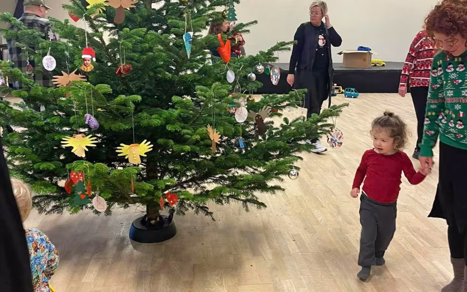 Børn og dagplejere samles omkring bordet, iført nissehuer, for at nyde et hyggeligt måltid sammen. Et øjeblik af fællesskab og julestemning