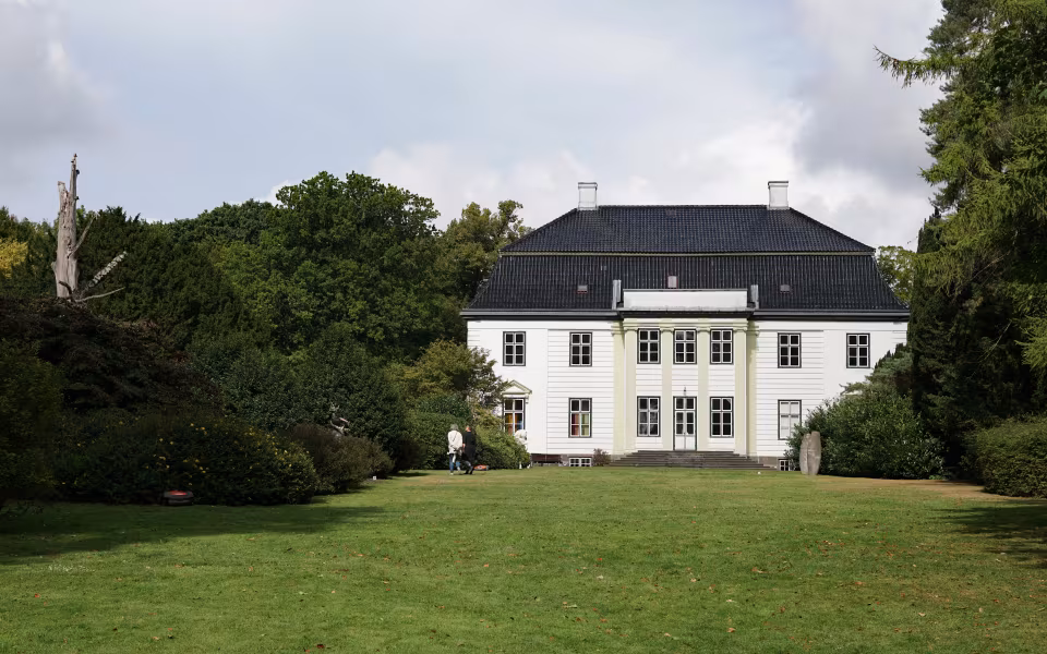 Hvid bygning ved Augustiana Kunstpark bag græsplæne og høje træer
