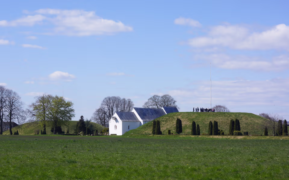 Jelling Kirke mellem gravhøje med besøgende på højderyggen ved Vikingernes Jelling