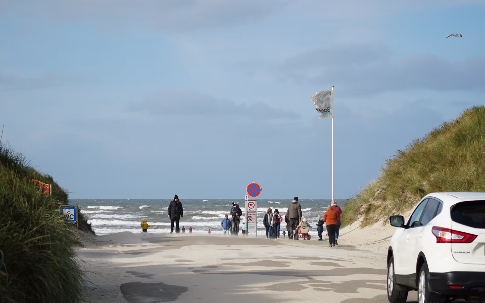 Sanddækket vej gennem klitter til Vejers Strand med biler og gående ved havet