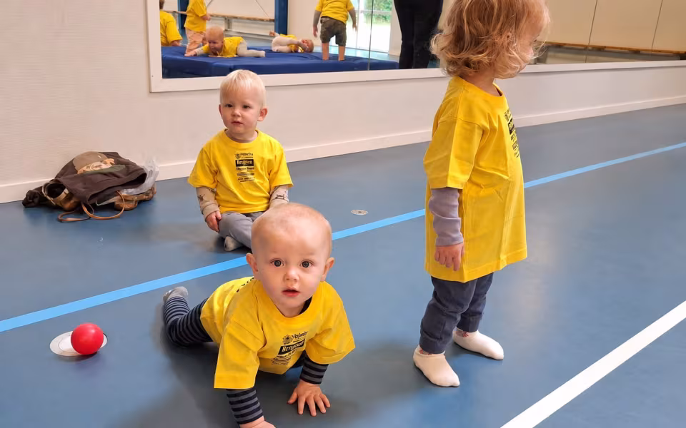 Dagplejere og børn mødes til bevægelse i Skovlund-Ansager Hallen, hvor de nye gule trøjer er i brug blandt farverige redskaber.