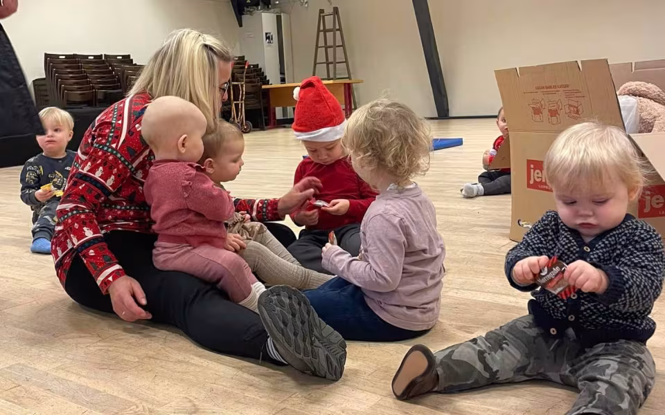 Børn og dagplejere samles omkring bordet, iført nissehuer, for at nyde et hyggeligt måltid sammen. Et øjeblik af fællesskab og julestemning