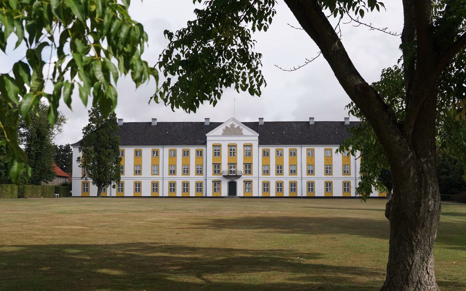 Augustenborg Slot med gul-hvid facade set bag store træer på plænen
