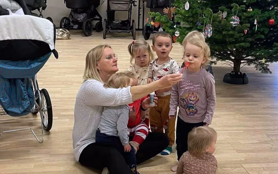 Børn og dagplejere samles omkring bordet, iført nissehuer, for at nyde et hyggeligt måltid sammen. Et øjeblik af fællesskab og julestemning