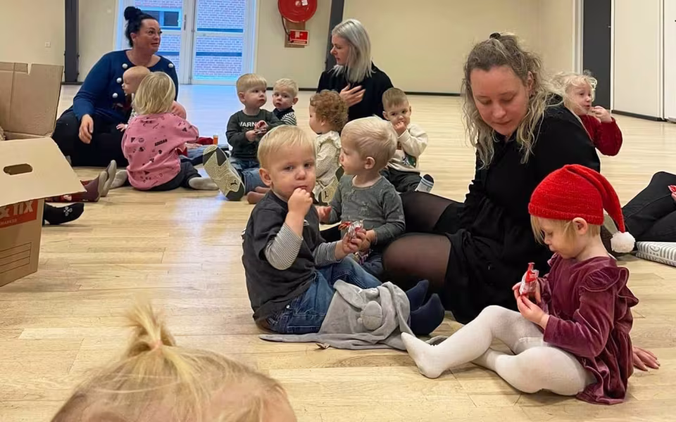 Børn og dagplejere samles omkring bordet, iført nissehuer, for at nyde et hyggeligt måltid sammen. Et øjeblik af fællesskab og julestemning