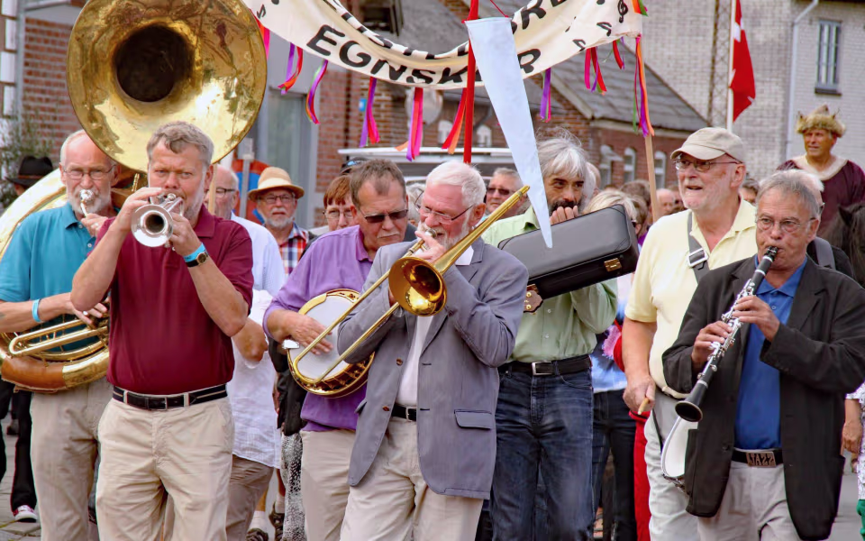 Brassband spiller i optog ved Mariefestival med faner og publikum i gaden