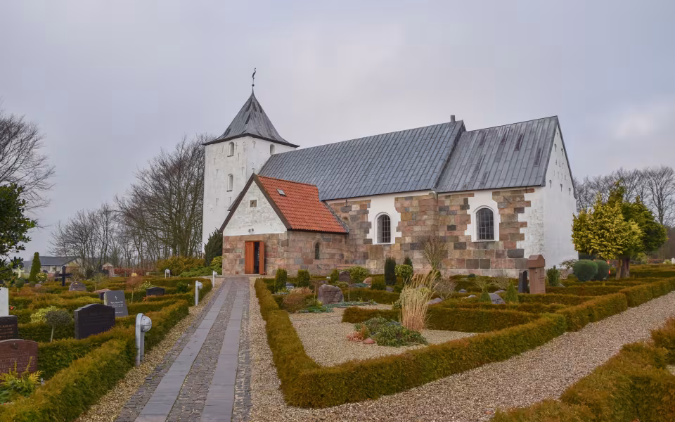 Sti gennem kirkegården mod Tistrup Kirkes hvide tårn og grå metaltag