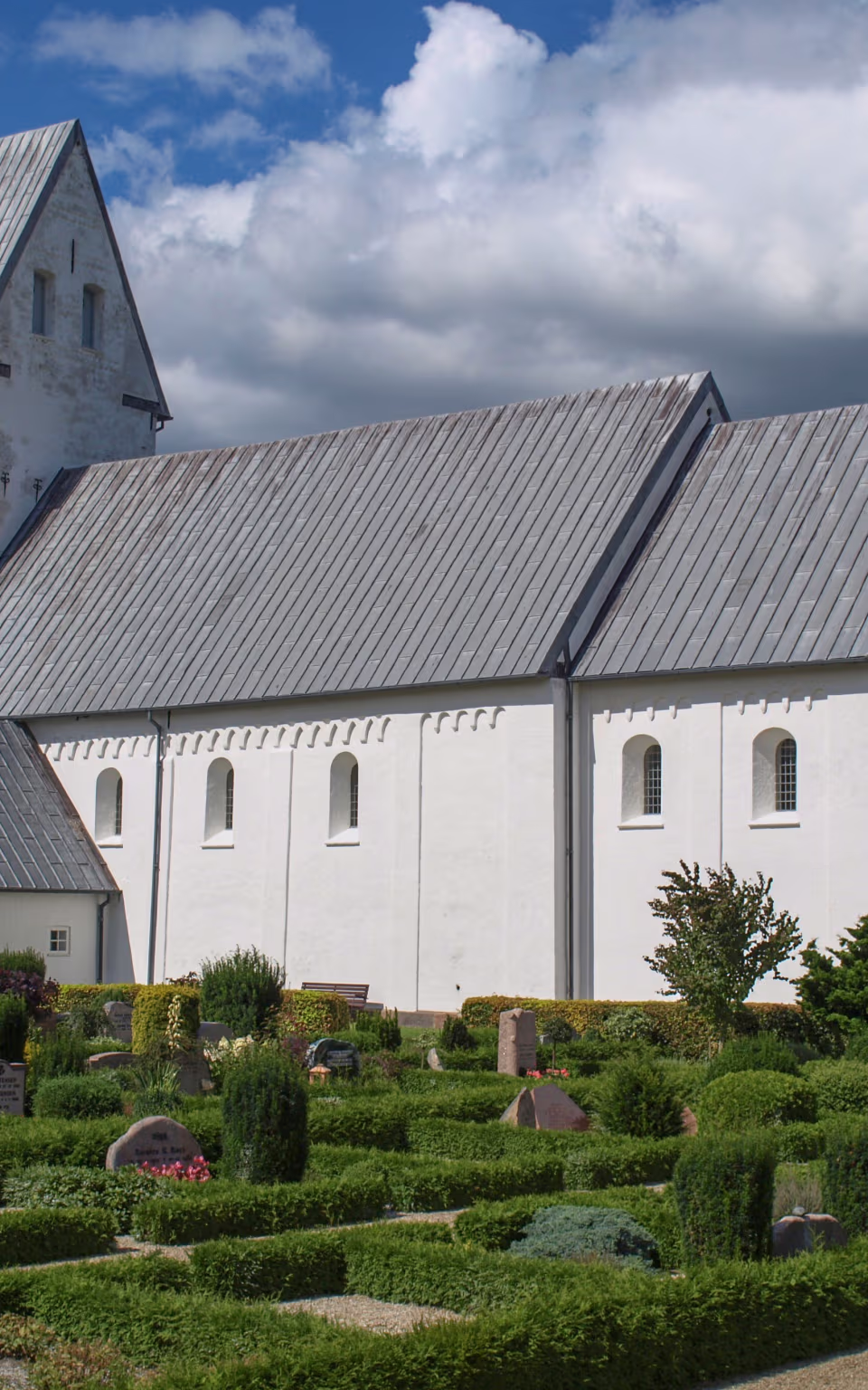 Billum Kirkes hvide tårn og kirkegård med hække under skyet blå himmel