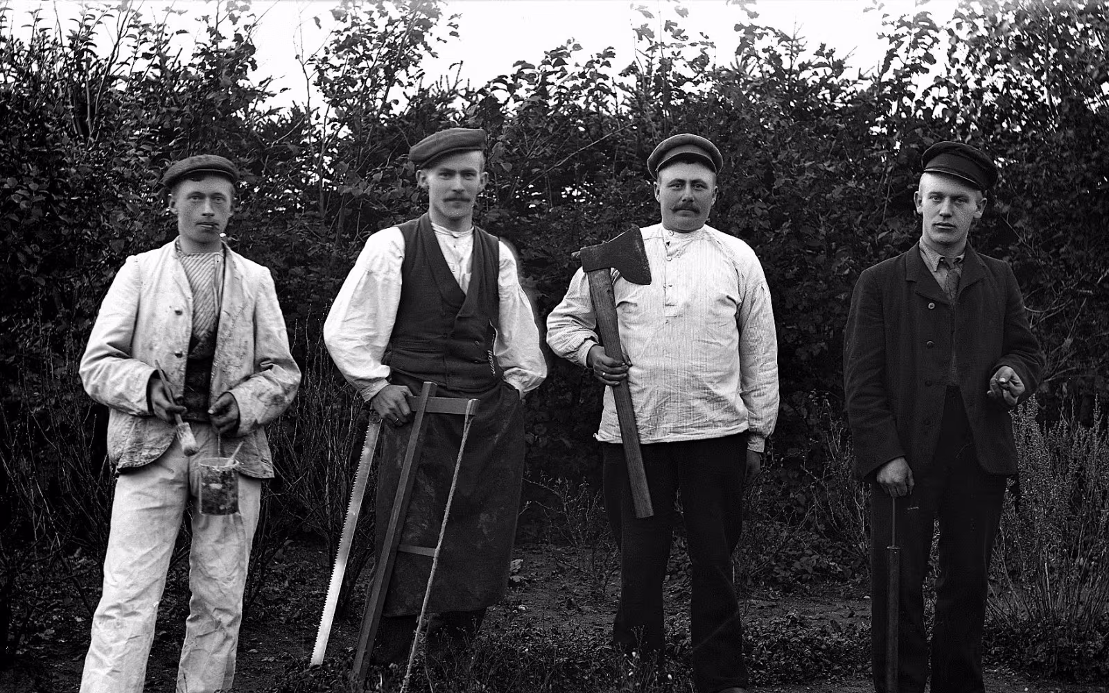 Fire mænd i Skovlund poserer med arbejdsredskaber foran tæt bevoksning