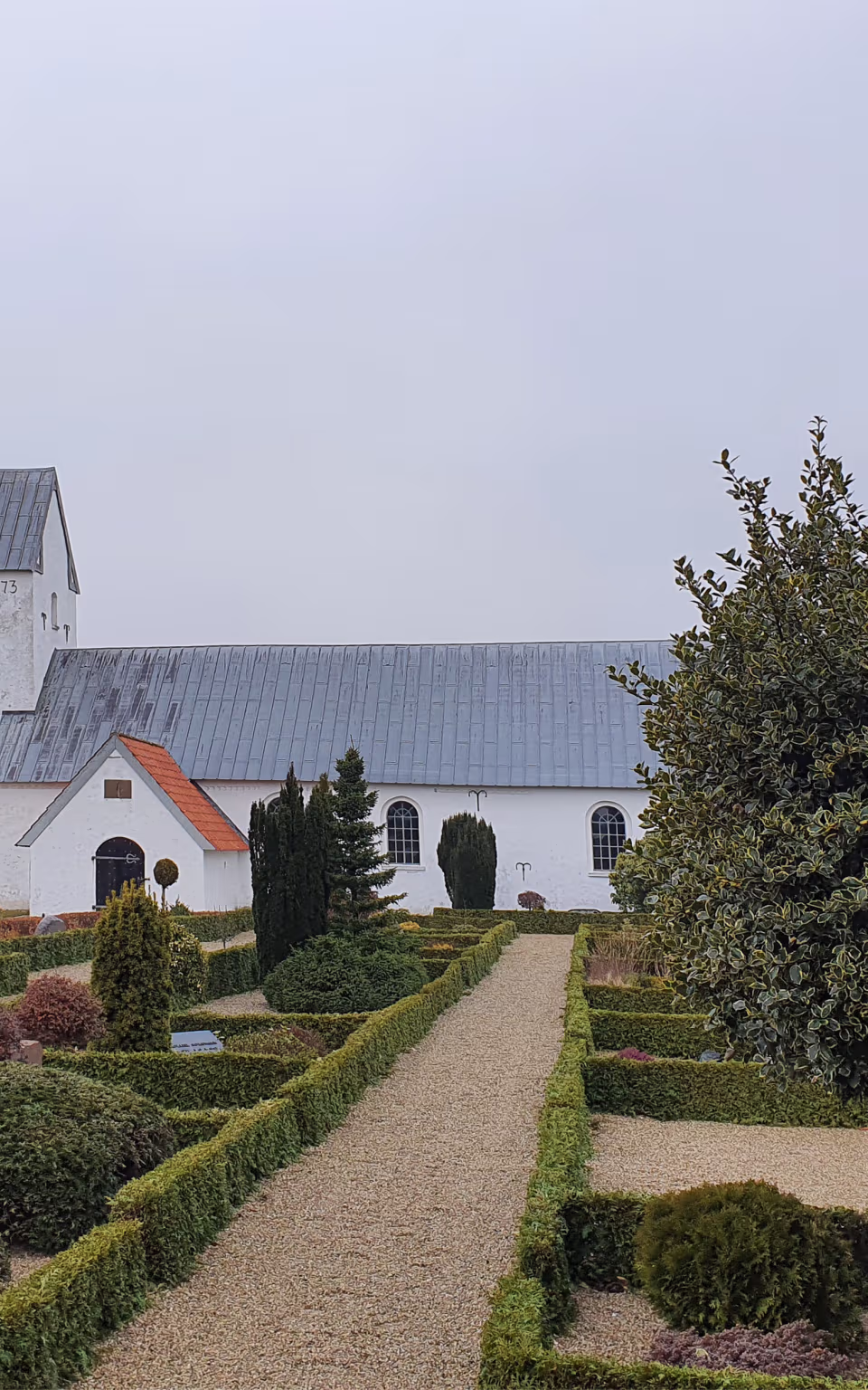 Kirke med hvidt tårn bag kirkegårdens hække og grussti