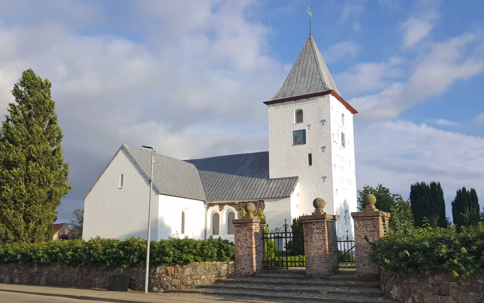 Hvid kirke med tårn, kirkegårdsmur og grøn hæk under blå himmel