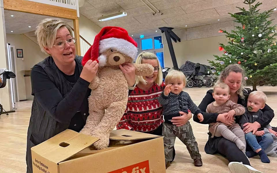 Børn og dagplejere samles omkring bordet, iført nissehuer, for at nyde et hyggeligt måltid sammen. Et øjeblik af fællesskab og julestemning