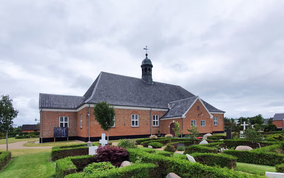 Kirkegård med hække og gravsten ved Nordby Kirke under overskyet himmel