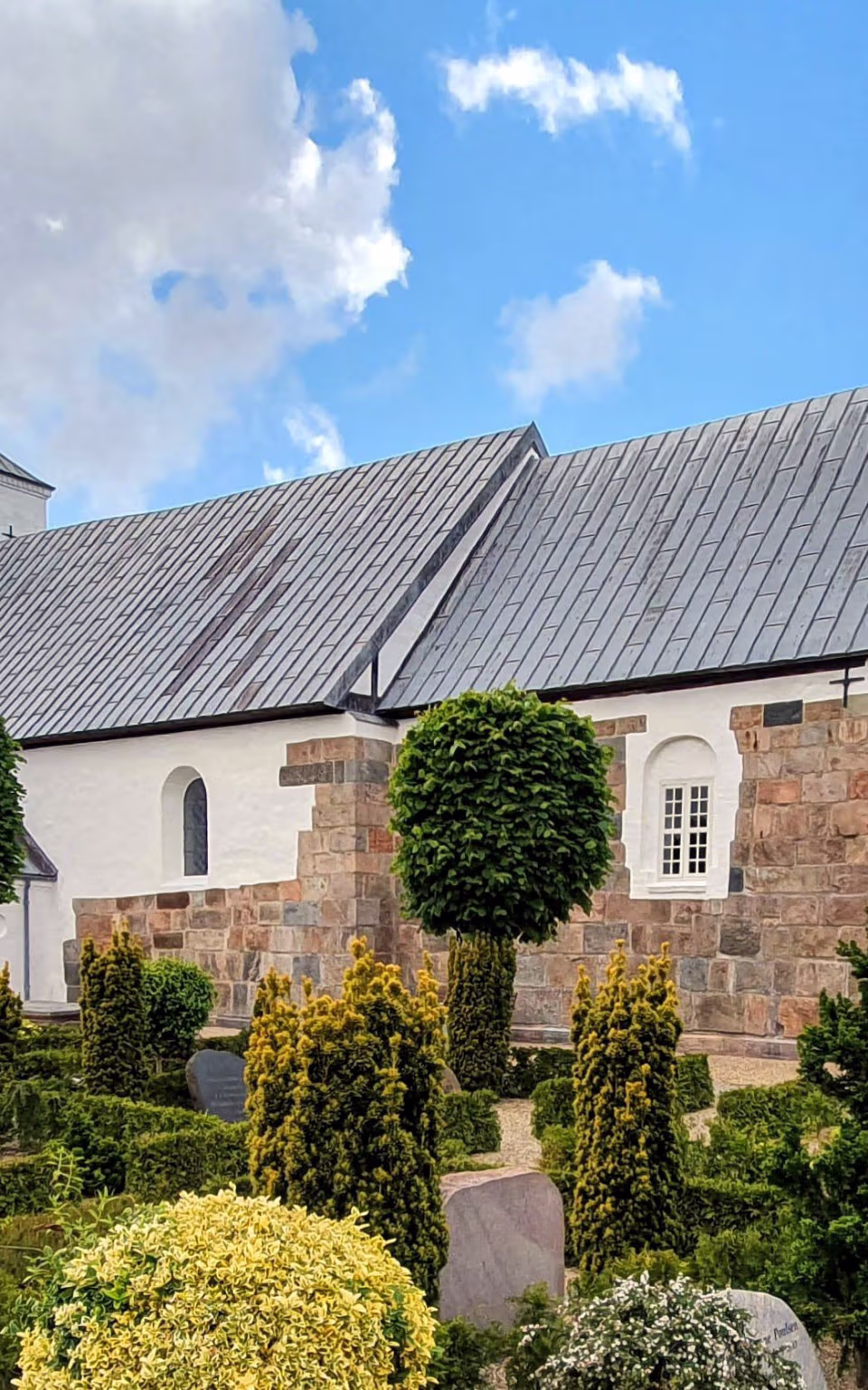 Outrup Kirke med kirkegård, klippede træer og gravsten under blå himmel