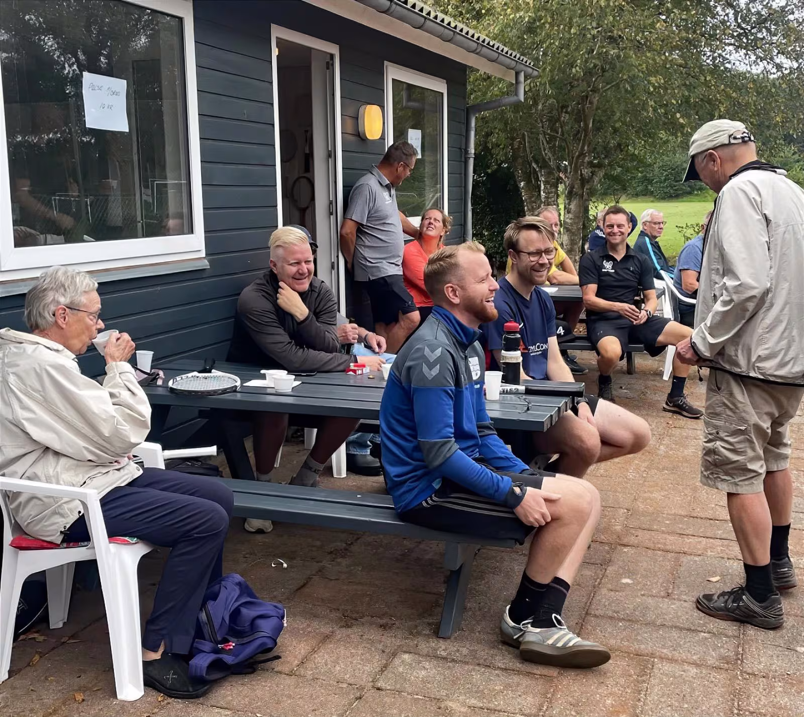 Glade Ansager Tennis Klub medlemmer samlet ved bordene foran klubhuset, midt i den årlige klubturnering. 75-års jubilæumsfesten venter senere på dagen. Tak til alle for en uforglemmelig dag!