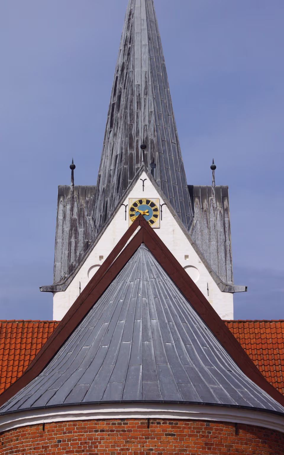 Kirketårn med gråt spir, ur og røde tegltage under blå himmel