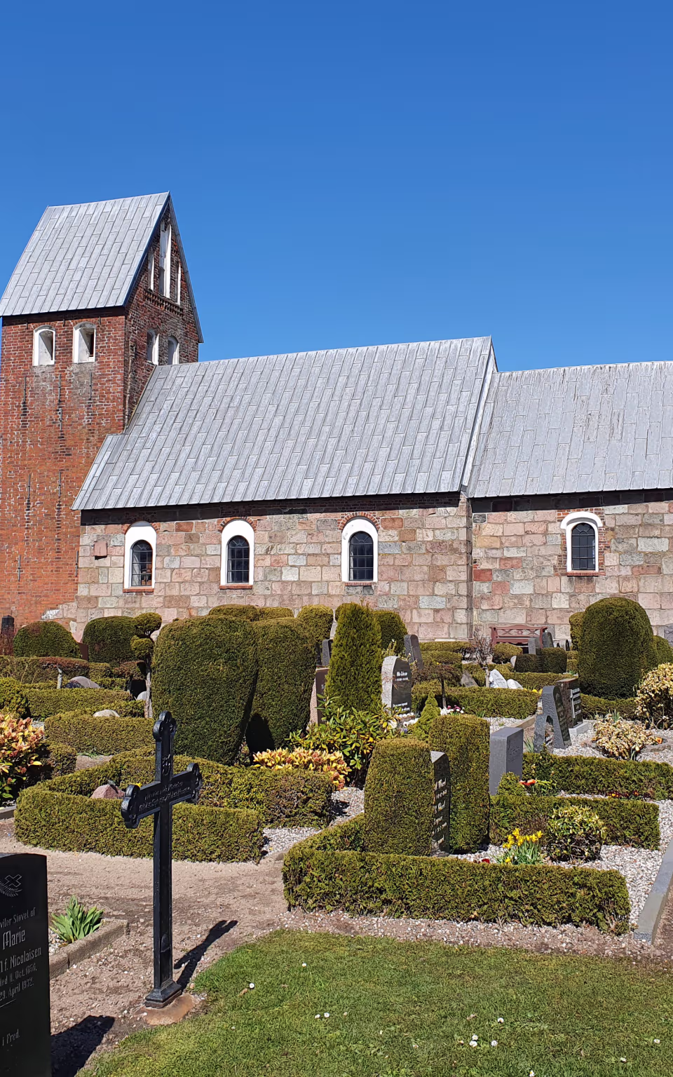 Hjerpsted Kirke med kirkegård, gravsten og klippede hække under blå himmel
