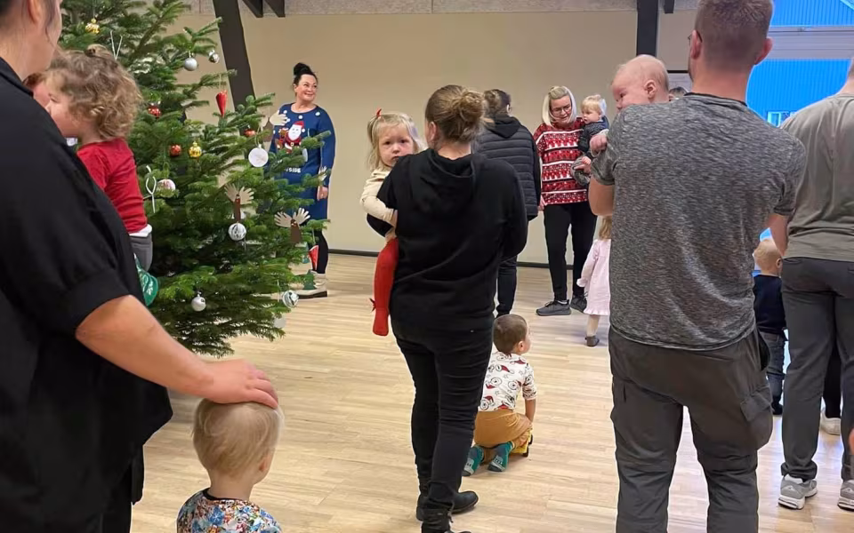 Børn og dagplejere samles omkring bordet, iført nissehuer, for at nyde et hyggeligt måltid sammen. Et øjeblik af fællesskab og julestemning