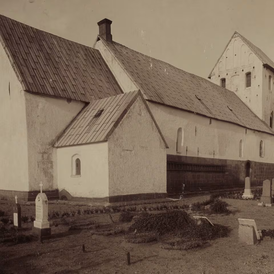 Janderup Kirke og kirkegården i 1906, tre år før den voldsomme stormflod, hvor omkring halvdelen af gravene sank sammen, og gravsten væltede. På kirkens sydside ses det gamle våbenhus, som blev revet ned i 1916. Foto: Kunstakademiets Bibliotek, 1906.