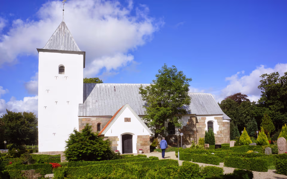 Hvidt kirketårn ved Thorstrup Kirke med kirkegård og grønne hække foran