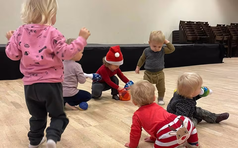Børn og dagplejere samles omkring bordet, iført nissehuer, for at nyde et hyggeligt måltid sammen. Et øjeblik af fællesskab og julestemning