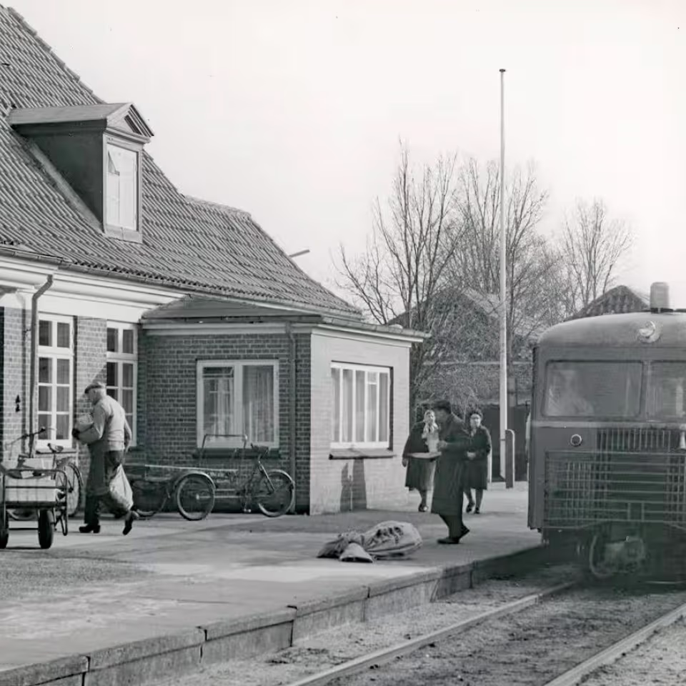 Foto fra togudstillingen: Persontoget fra Grindsted er netop ankommet til Ansager Station, hvor post og pakker bliver fordelt. I forgrunden ses Hans S. Nissen, kaldet “Pakhans”, som kom cyklende fra byen på sin gamle budcykel. Han kørte gennem mange år rundt med pakker i Ansager – først med hestevogn, senere på cykel