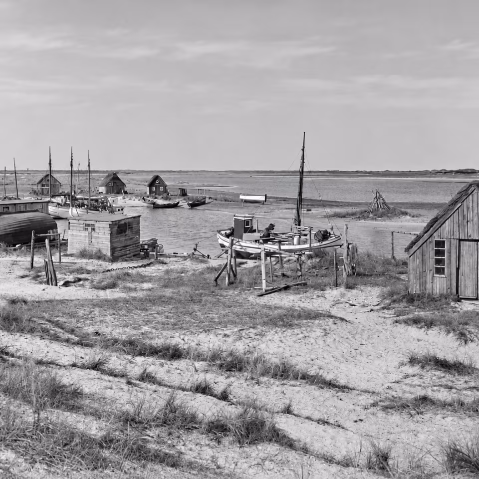 Foto: Achton Friis, 1923. Nymindegabs gamle bådehavn set mod nord. Masterne rager op mellem de små fiskerhytter og træskure, mens bådene ligger fortøjet eller er trukket på land. Overalt ses spor af fiskernes daglige arbejde. Fotografiet er et tidsbillede fra det gamle fiskerleje og indgår i Achton Friis' omfattende dokumentation af danske landskaber og levevilkår. Kilde: Det Kgl. Biblioteks Billedsamling.