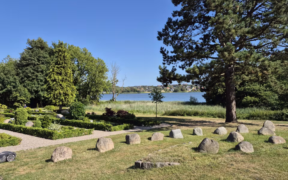 Gravsten og grønne træer på Damager Kirkegård med vand i baggrunden