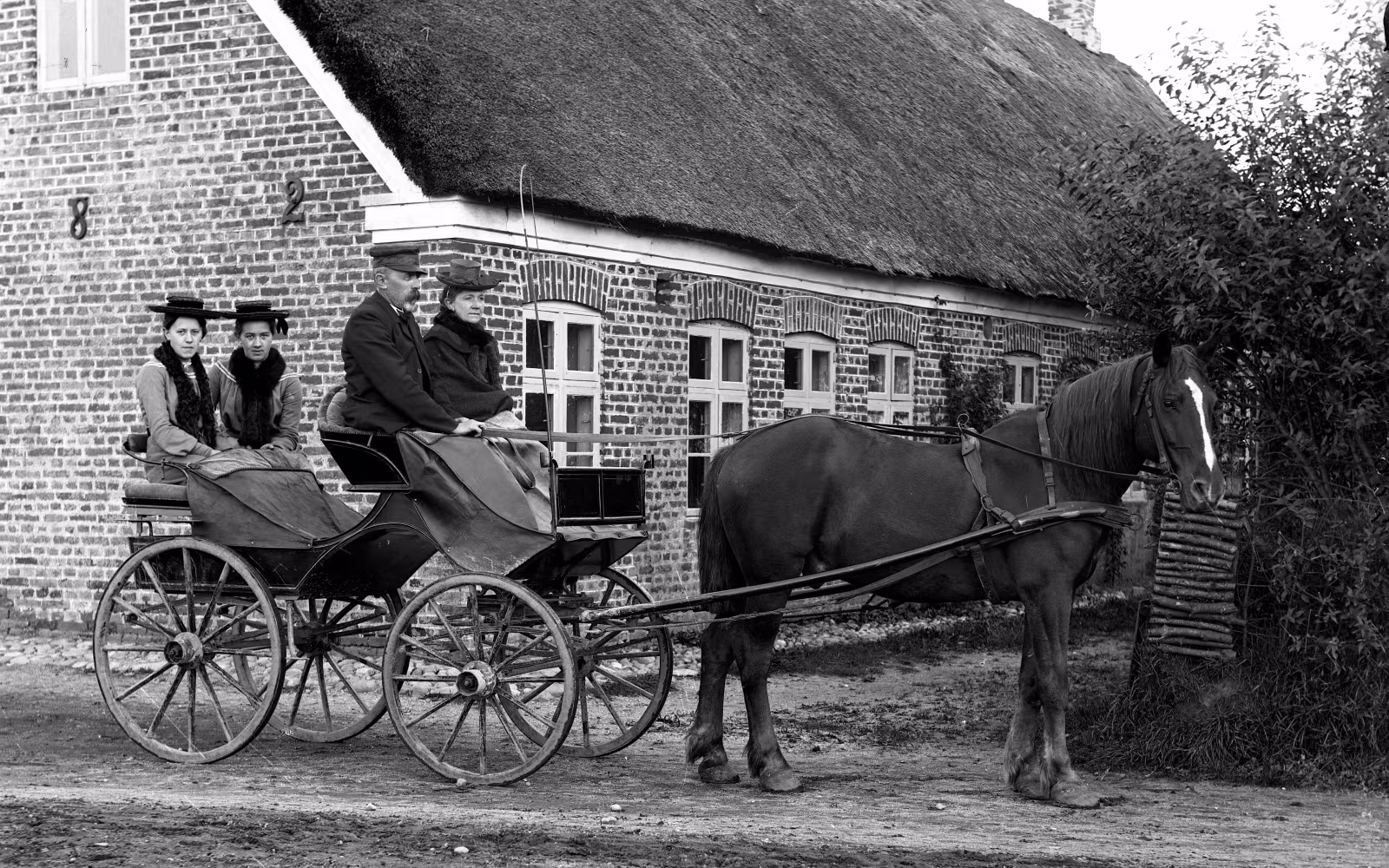 Hestevogne med passagerer foran stråtækt murstenshus i Skovlund