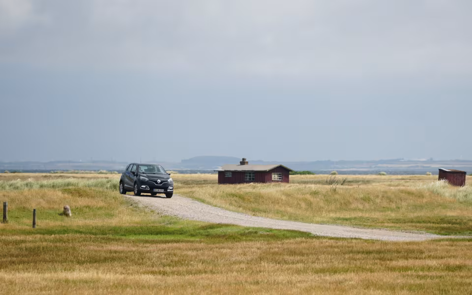 Sort bil på grusvej ved Gjellerodde mellem sommergræs og små hytter