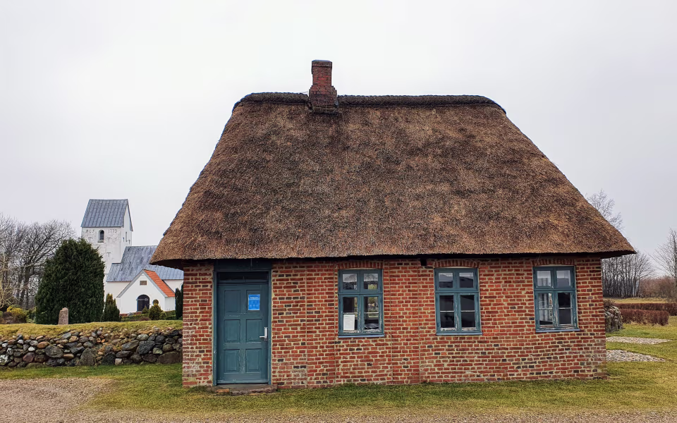 Stråtækt murstensbygning ved Hodde Gamle Skole med kirketårn i baggrunden