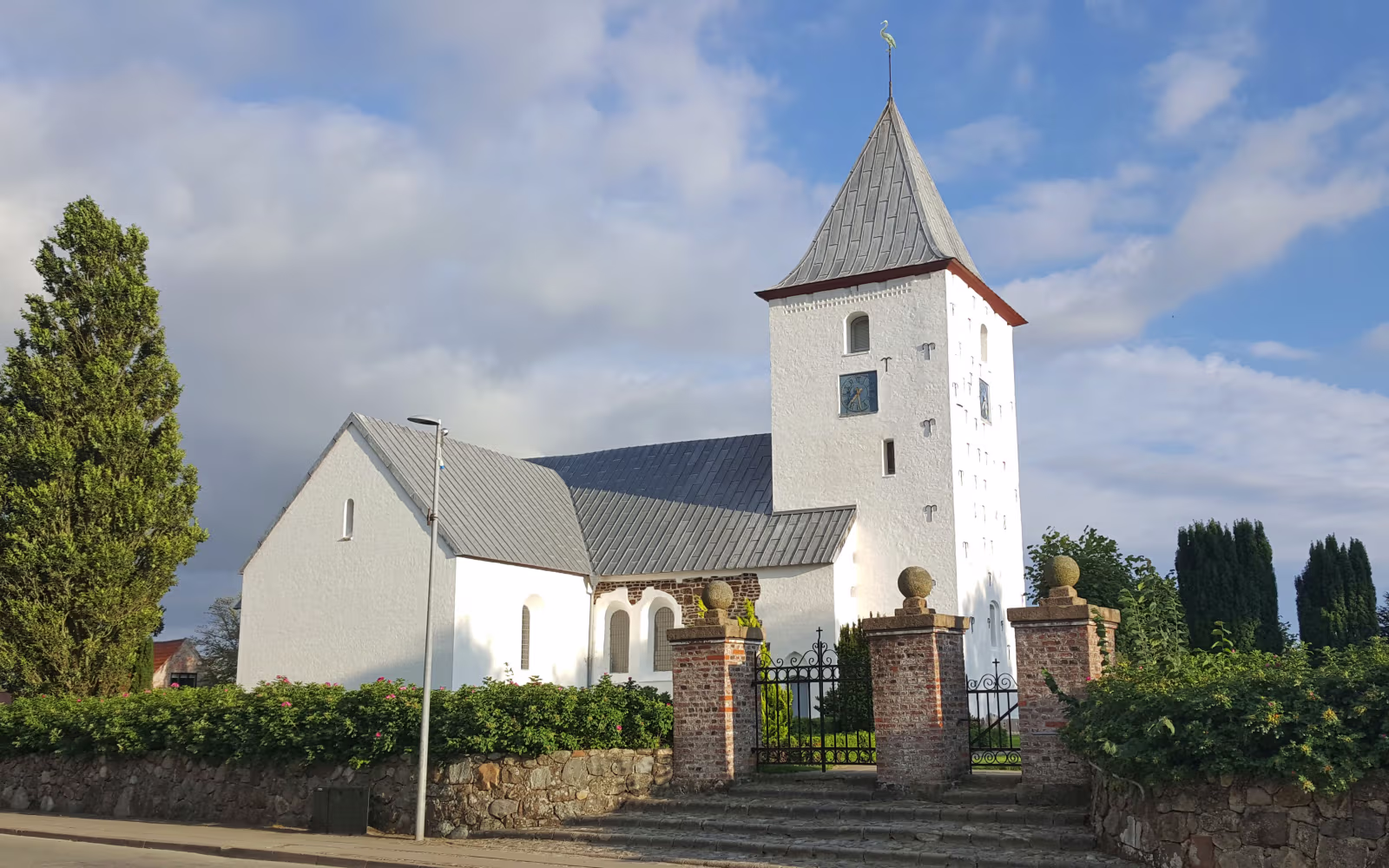 Hvid kirke med tårn, kirkegårdsmur og grøn hæk under blå himmel