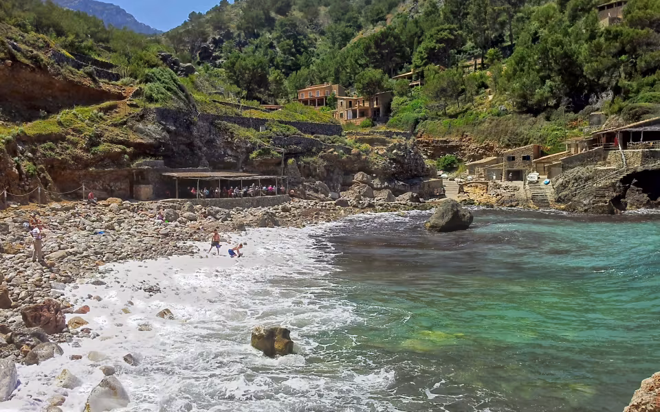 Klippestrand med badende ved Cala Deià under terrasserede huse og fyrretræer