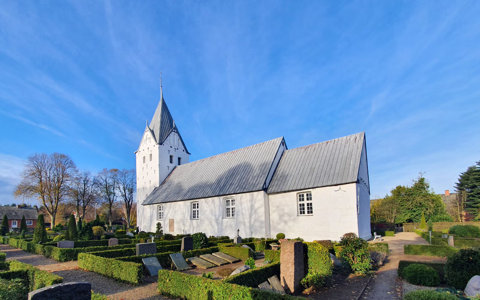 Arrild Kirke med hvidt tårn over kirkegård med hække og gravsten