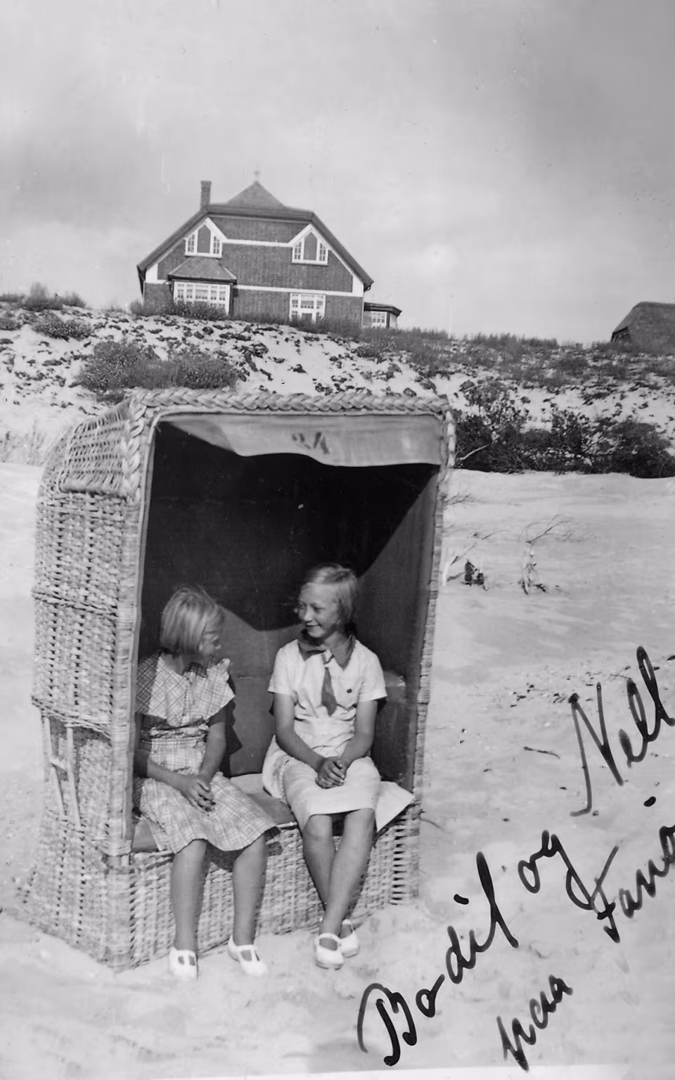 To piger sidder i strandkurv foran klitter og hus på udflugt fra Skovlund