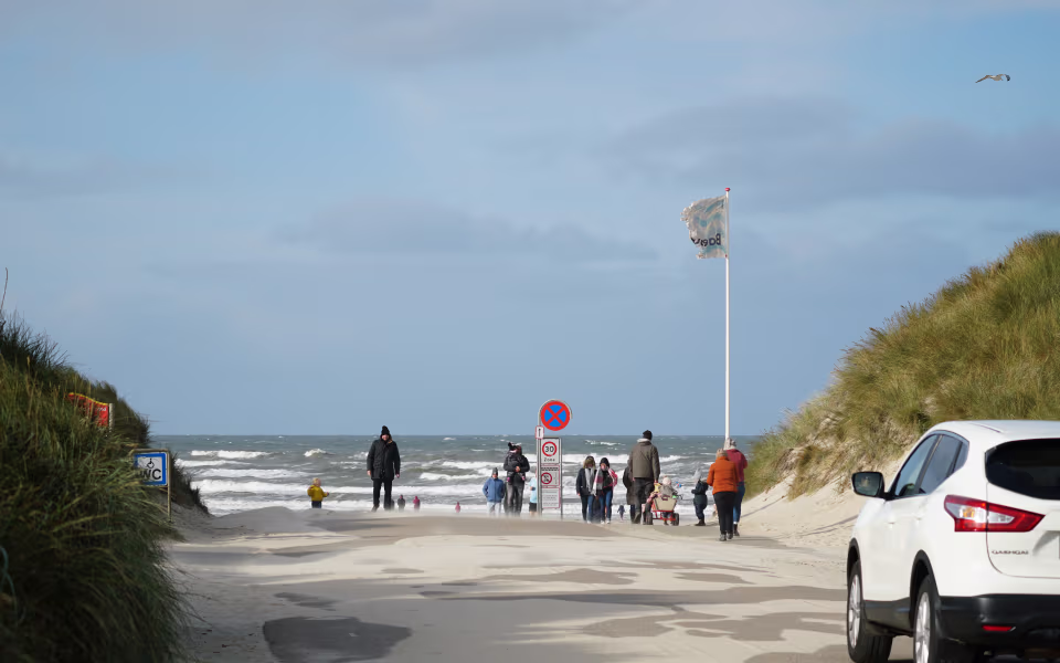 Sanddækket vej gennem klitter til Vejers Strand med biler og gående ved havet