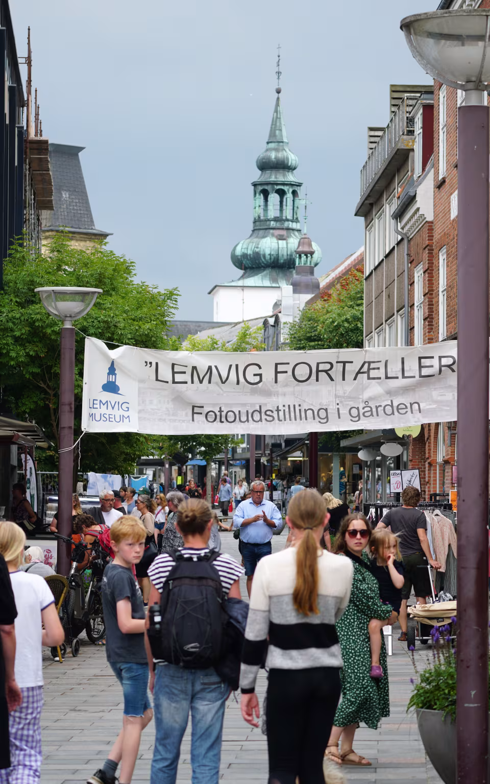 Travl gågade i Lemvig med shoppende mennesker, museumsbanner og kirketårn i baggrunden