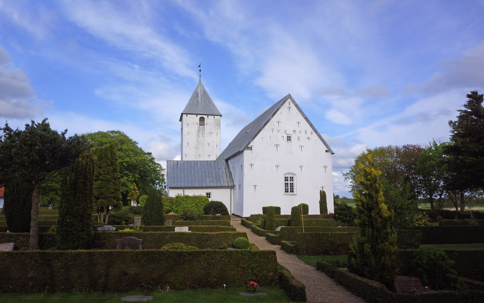 Vilslev Kirke med hvidt tårn og kirkegård med hække under blå himmel