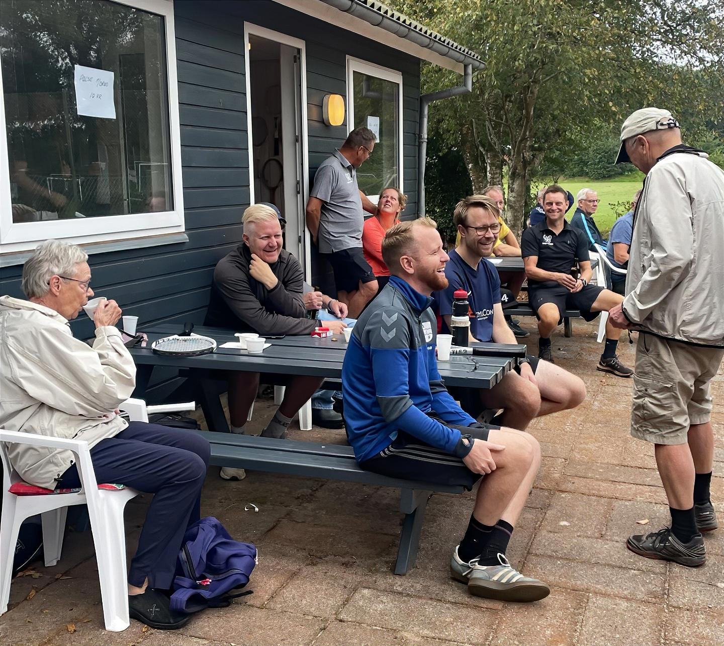 Glade Ansager Tennis Klub medlemmer samlet ved bordene foran klubhuset, midt i den årlige klubturnering. 75-års jubilæumsfesten venter senere på dagen. Tak til alle for en uforglemmelig dag!