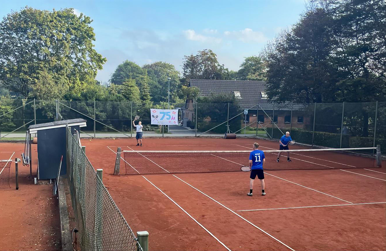 Glade Ansager Tennis Klub medlemmer samlet ved bordene foran klubhuset, midt i den årlige klubturnering. 75-års jubilæumsfesten venter senere på dagen. Tak til alle for en uforglemmelig dag!