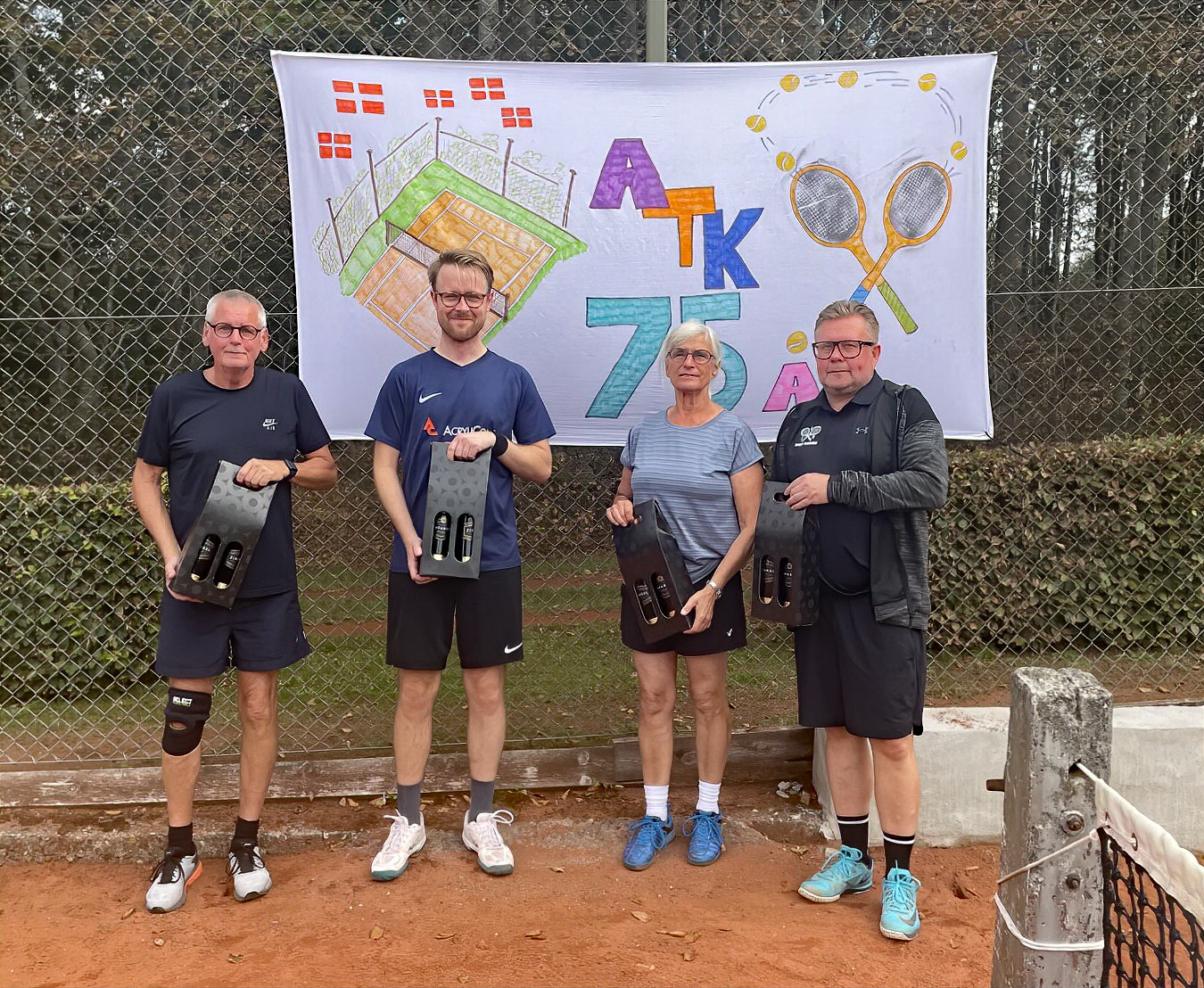 Glade Ansager Tennis Klub medlemmer samlet ved bordene foran klubhuset, midt i den årlige klubturnering. 75-års jubilæumsfesten venter senere på dagen. Tak til alle for en uforglemmelig dag!